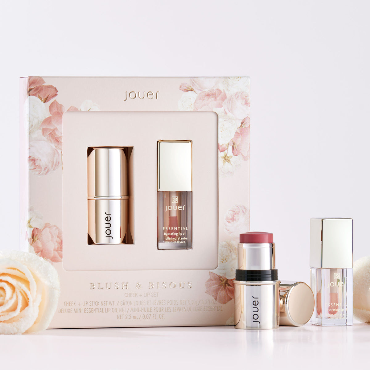 A boxed set from Jouer Cosmetics, labeled "Blush & Bisous," is displayed against a light background adorned with floral accents. This cruelty-free set features a rose gold blush stick and a clear lip gloss, both arranged neatly amidst additional roses.