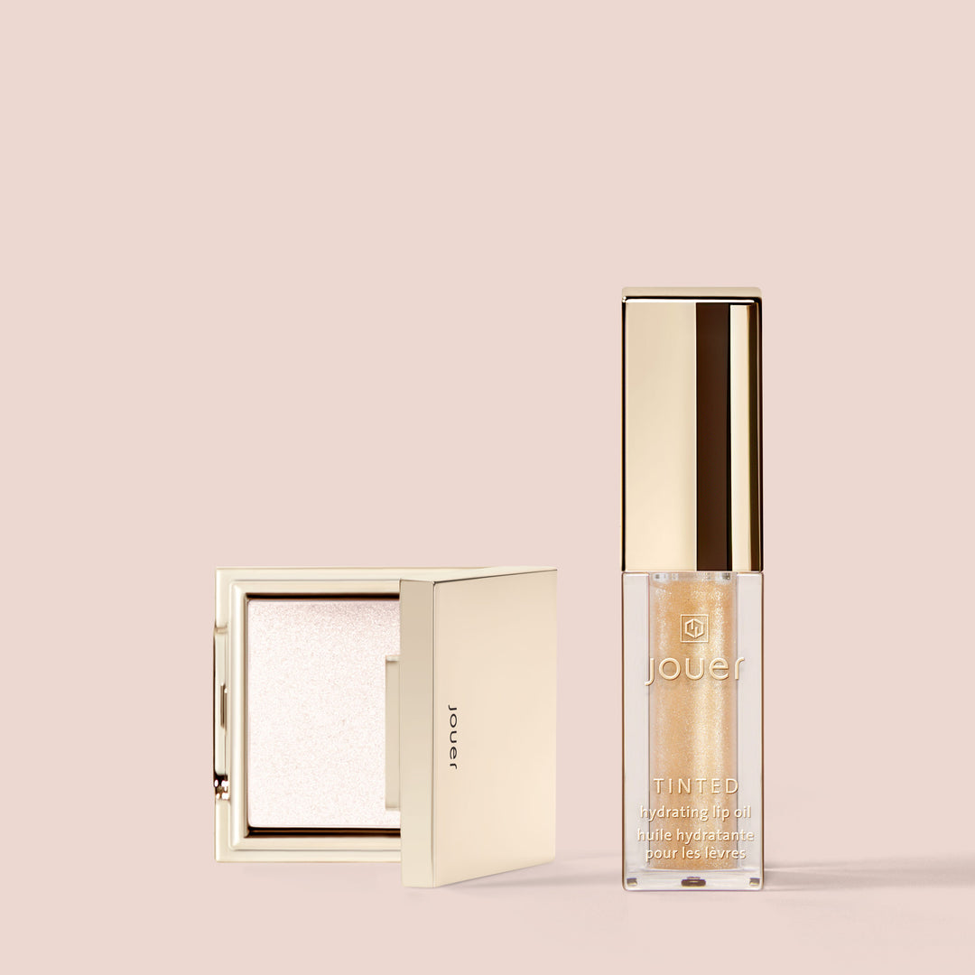 A minimalistic beige background showcases two makeup products: Jouer Cosmetics' Glow-on La Go, featuring a square compact with a reflective off-white highlighter on the left and a rectangular tube of paraben-free tinted hydrating lip oil with a beige-gold cap on the right.