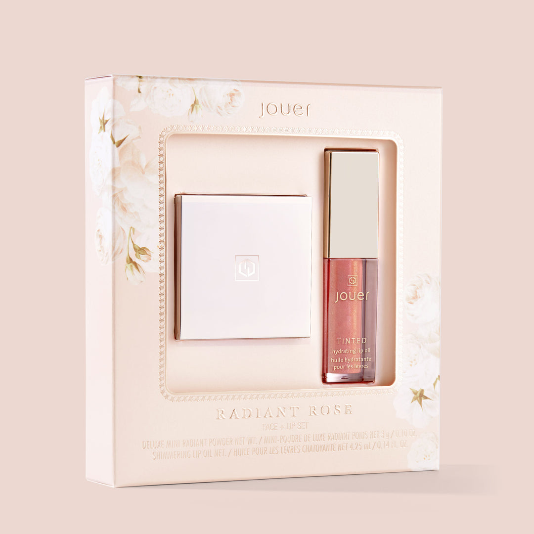 A makeup set titled "Radiant Rose" by Jouer Cosmetics comes in a beige-toned box adorned with floral accents on the left side. Inside, you'll find a compact of Radiant Translucent Powder and a tube of shimmering lip oil, both featuring elegant gold caps. This cruelty-free set includes mini versions of the radiant powder and shimmering lip oil.