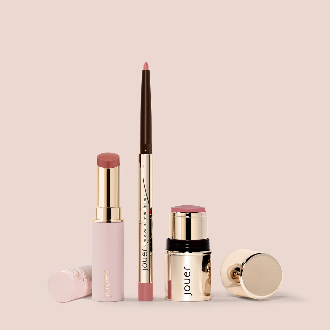 A collection of Jouer Cosmetics products against a beige background, featuring the Bare Rose set that includes a pink lipstick in a gold tube, a Shine Balm blush stick with a gold cap, and a brown lip liner with gold accents. The brand name 'Jouer Cosmetics' is elegantly displayed on each item.