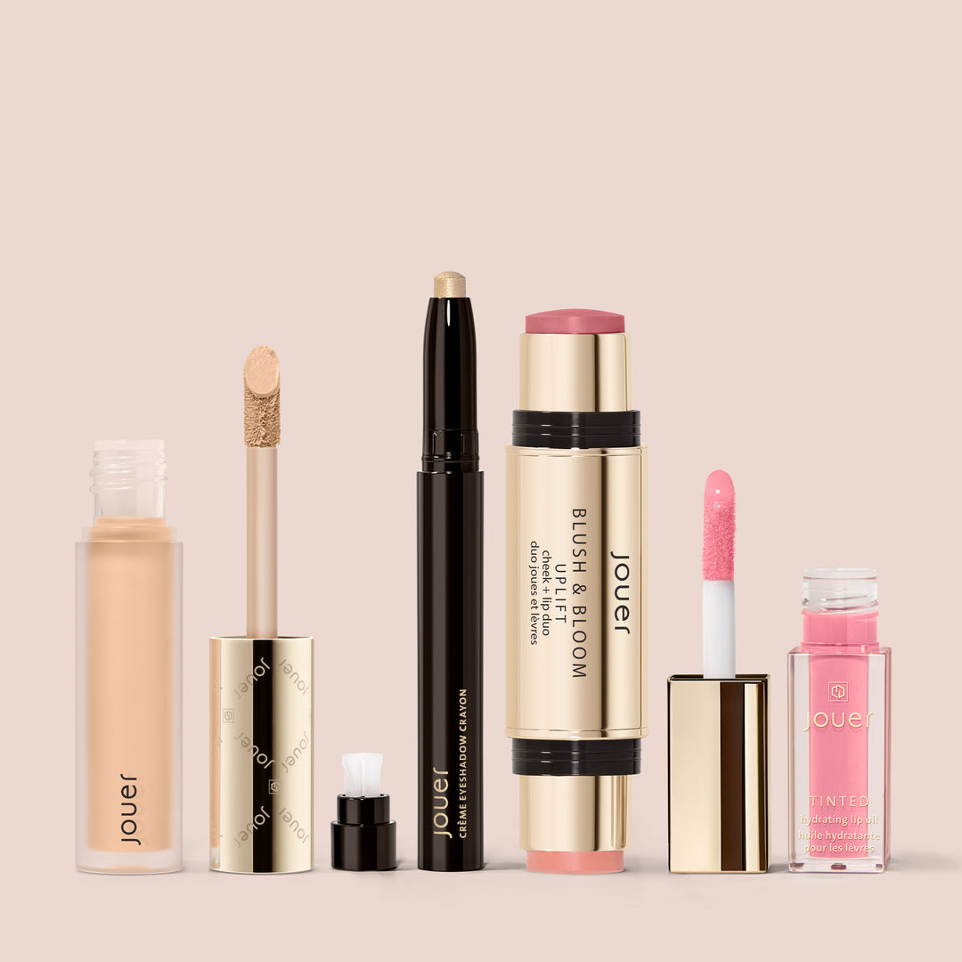 The French Girl Makeup Set by Jouer Cosmetics displayed against a neutral beige background features an array of products, including a liquid concealer, highlighter stick, blush and bloom stick, and a tinted lip oil in a clear tube with a pink cap. Each item is elegantly packaged in gold and clear containers.