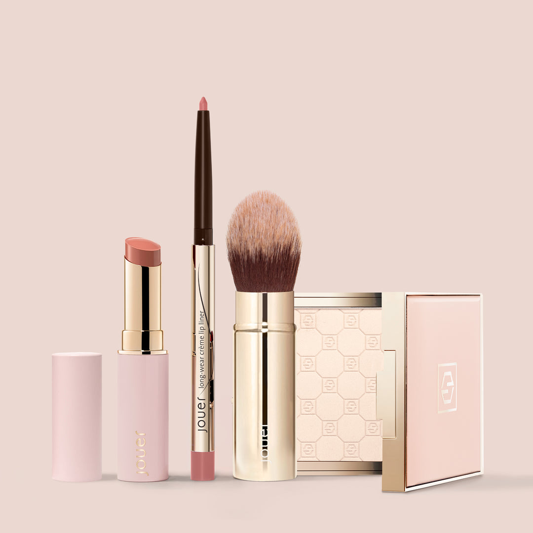 The On-The-Go Set by Jouer Cosmetics, featuring a lipstick, lip liner, makeup brush, and compact case with mirror in elegant minimalist designs, is neatly arranged against a matching light pink background. This collection of cruelty-free, light pink clean beauty cosmetics provides both style and substance.