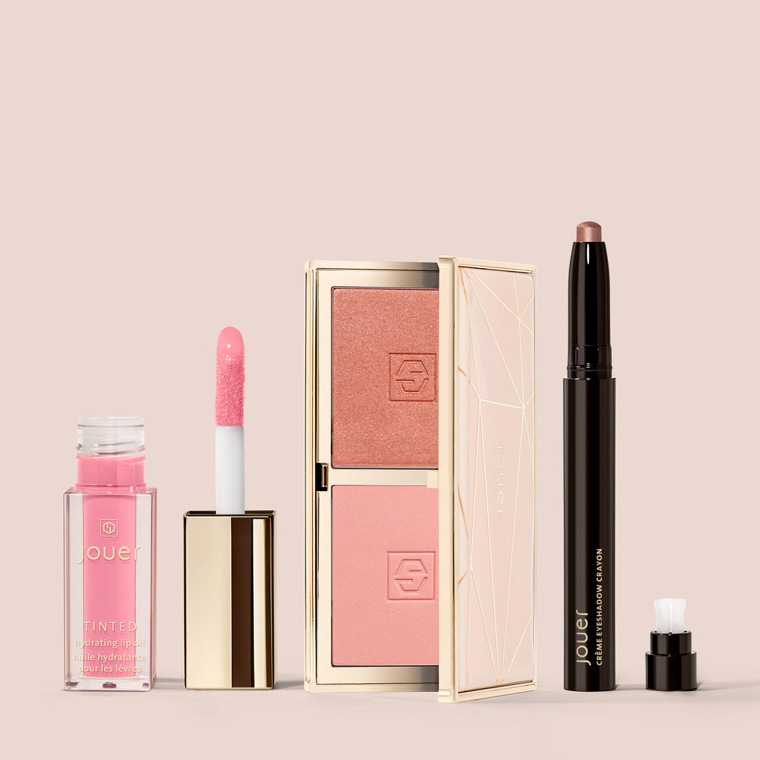 A set of beauty products by Jouer Cosmetics against a beige background. From left to right: a small bottle of pink Tinted Lip Oil with an open applicator, a compact case with two shades from the Le Blush Set, and a Crème Eyeshadow Crayon in a sleek black and gold tube with the cap off.