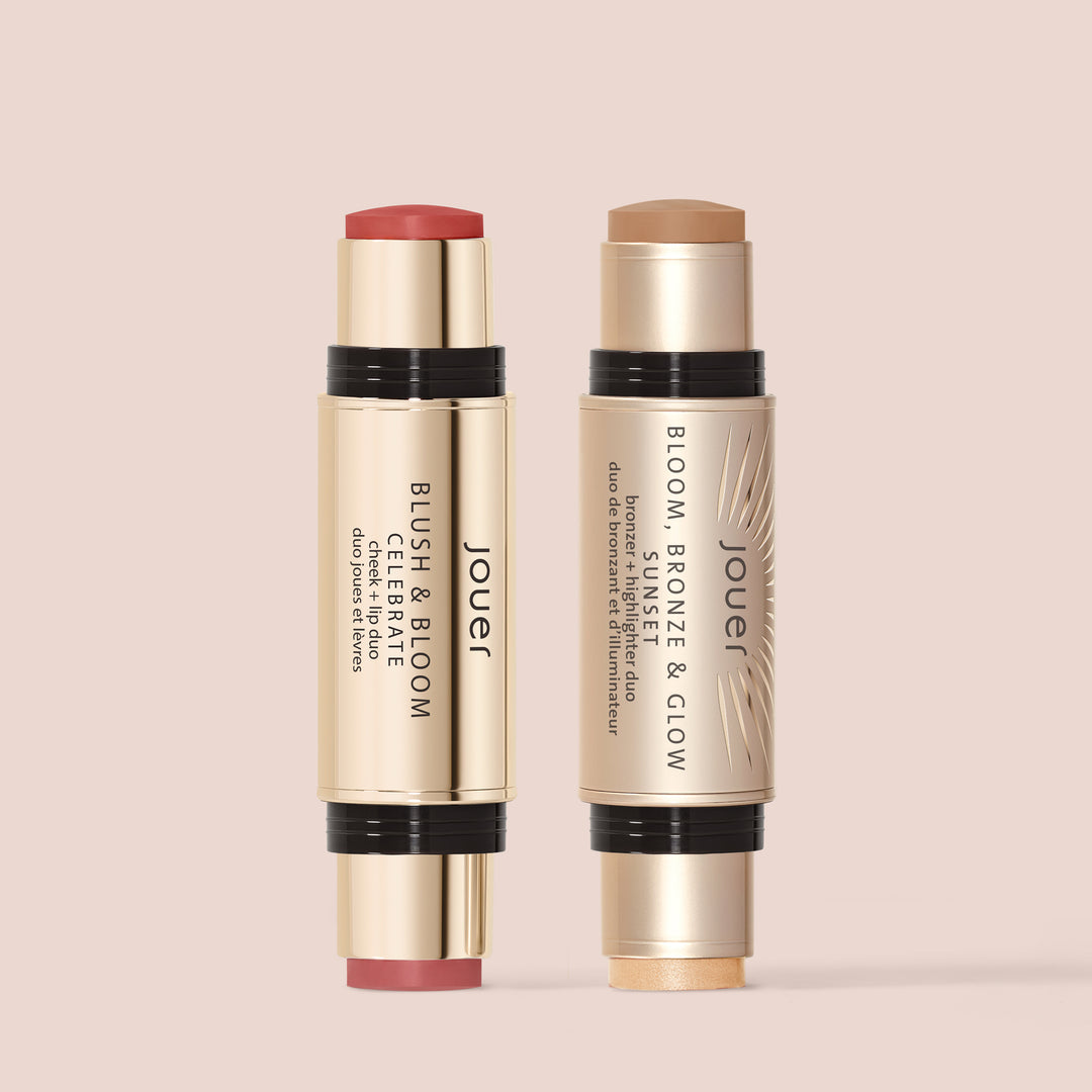 Two Jouer Cosmetics beauty sticks from Le Cheek Set stand against a peach background. One dual-ended stick is labeled "Blush & Bloom Duo" with blush and highlighter ends, while the other is labeled "Bloom Bronze & Glow Duo," featuring bronzer and highlighter ends. Both products boast sleek, gold packaging and are certified cruelty-free.