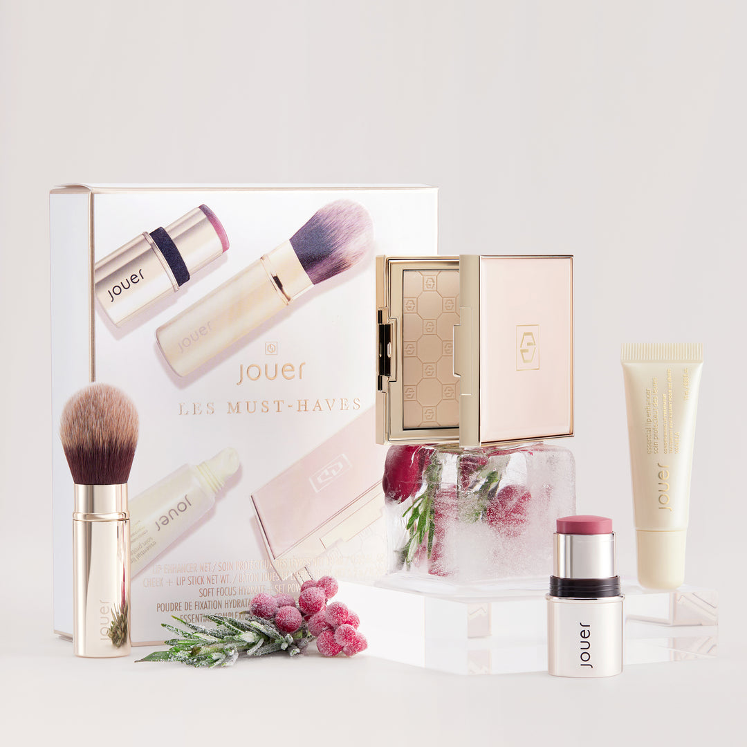 Introducing Les Must-Haves by Jouer Cosmetics: a luxurious makeup set featuring a gluten-free lipstick, blush stick, foundation brush, compact case, and face primer. This elegant collection is beautifully packaged in gold and beige with frosty accents and adorned with small berry decorations to complement the main items.