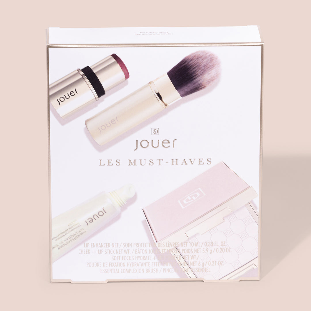 The image displays the "Les Must-Haves" boxed set of beauty products by Jouer Cosmetics. This collection features a lip enhancer, a cheek and lip stick, a foundation primer, and a complexion brush, all elegantly presented within the box. The sleek packaging showcases a minimalistic design while emphasizing that the products are cruelty-free and gluten-free.