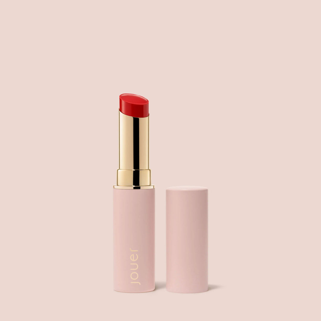 Karma, A tube of warm red, vegan "Balm Bouche" lipstick by Jouer Cosmetics, featuring a light pink casing with gold accents and branded with "Jouer" near the base. The tube is open to reveal the gluten-free lipstick, with the cap placed beside it. The background is a solid light pink color.