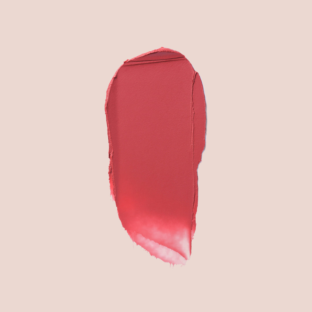 Solidarite, A swatch of Balm Bouche from Jouer Cosmetics in the shade Solidarite smeared on a light pink background. The texture exhibits a smooth and creamy consistency, with the color transitioning from a medium muted rose shade at the top to a softer, lighter red towards the bottom. This vegan formula ensures both vibrant color and ethical beauty.