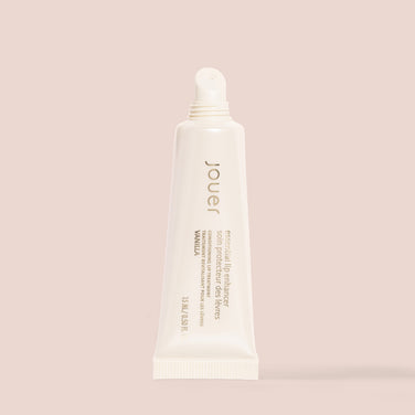  A tube of Jouer Cosmetics Essential Lip Enhancer, which is both cruelty-free and paraben-free, stands upright on a beige background. The off-white tube with a narrow nozzle displays text that identifies it as a conditioning lip treatment in vanilla flavor, containing 0.5 fl oz or 15 mL.