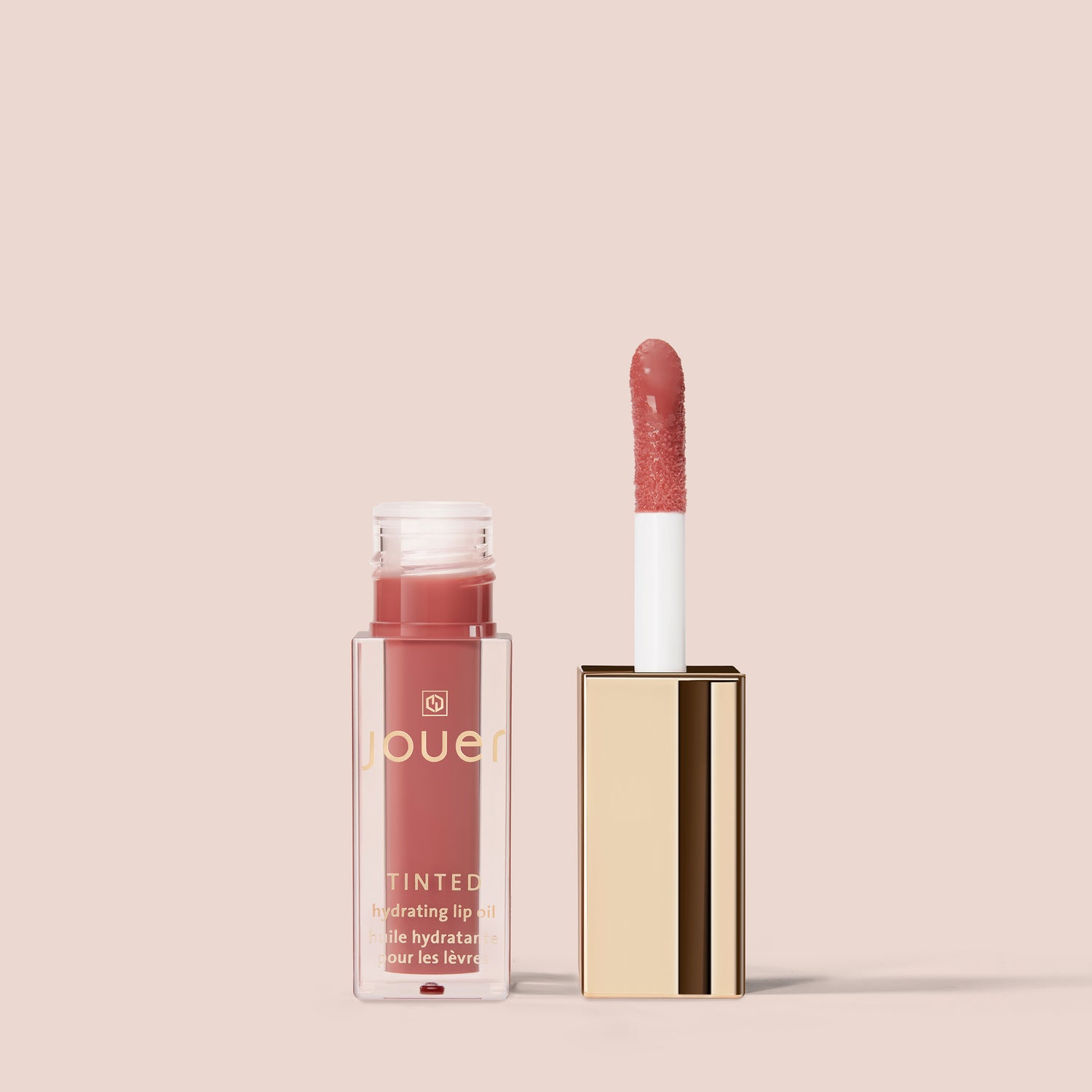 Bare rose, A tube of Tinted Lip Oil from Jouer Cosmetics, which is both vegan and paraben-free, is shown against a light pink background. The tube is open, revealing a pink-tinted lip oil with a white applicator stick and a gold cap placed next to the tube.