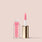 A pink-tinted, paraben-free lip gloss from Jouer Cosmetics in a clear rectangular tube with a gold cap. The cap is off, revealing a white wand with a pink applicator tip standing beside the tube on a beige background. The text on the tube reads "Jouer Tinted Lip Oil," proudly vegan and gluten free.