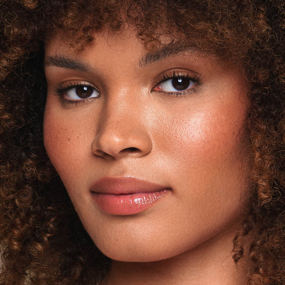 A close-up of a person's face showcases curly brown hair, clear skin, and glossy lips enhanced by Jouer Cosmetics' Essential Lip Enhancer. The lighting highlights their natural features and smooth complexion.