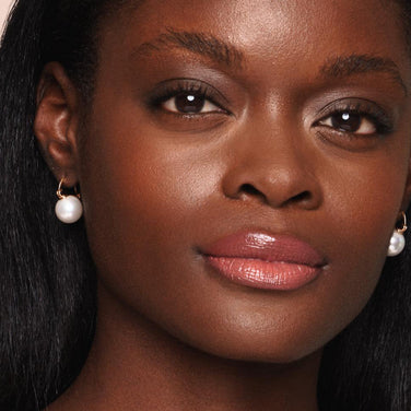  Close-up of a person with smooth, glowing dark skin. They are wearing shimmery eye shadow, glossy pink Essential Shine Balm by Jouer Cosmetics, and gold earrings with white pearls. The background is plain. Their radiant look is enhanced with cruelty-free products that align beauty with conscience.