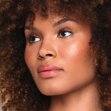  Close-up of a person with smooth, glowing dark skin. They are wearing shimmery eye shadow, glossy pink Essential Shine Balm by Jouer Cosmetics, and gold earrings with white pearls. The background is plain. Their radiant look is enhanced with cruelty-free products that align beauty with conscience.