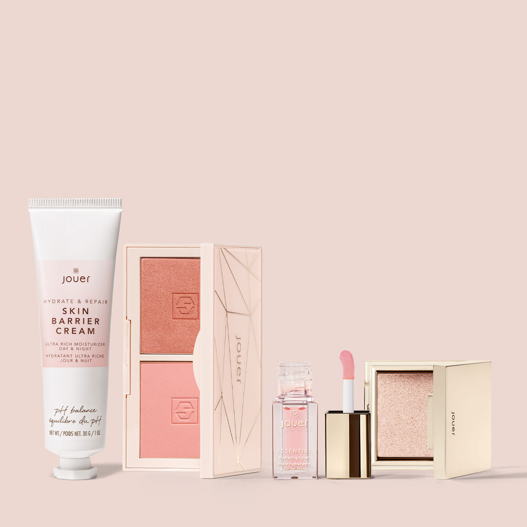 An arrangement of beauty products from Jouer Cosmetics' Mini Travel Set against a soft pink background. Included are a tube of Skin Barrier Cream on the left, a Mini Blush Bouquet, a clear Deluxe Mini Lip Oil, and a small square compact of shimmer highlighter.