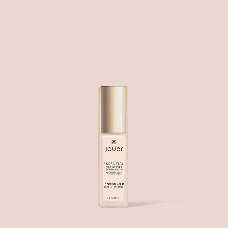 Jouer Essential high coverage creme foundation bottle with gold cap on beige background