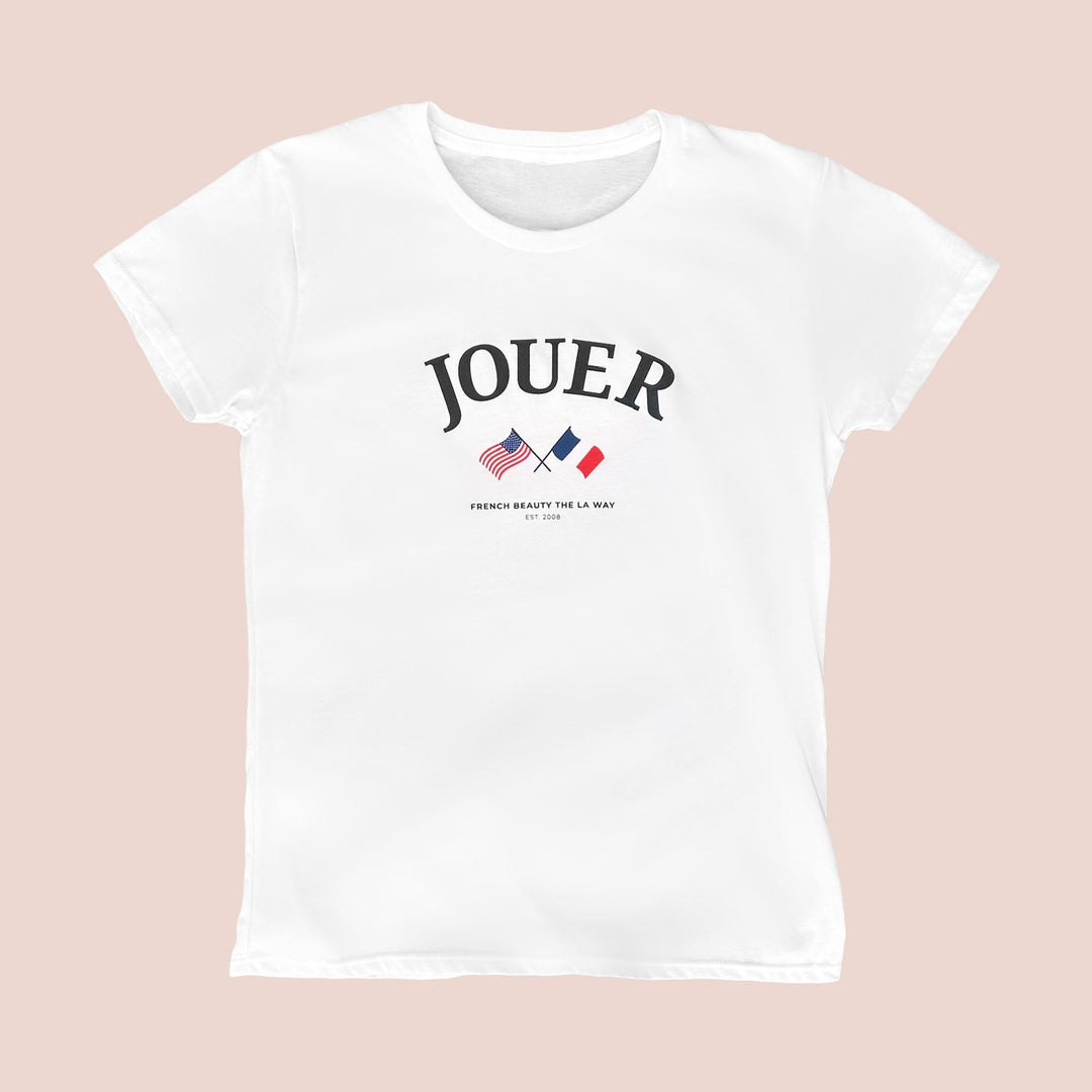 The Paris Meets LA Tee by Jouer Cosmetics is a white t-shirt crafted from 100% cotton. It features the word "Jouer" printed prominently in bold black letters at the chest, with an illustration of the American and French flags crossed below. Accompanying this design are the texts "French Beauty The LA Way" and "Est. 2008" underneath. The prewashed shirt has a light beige background and is machine washable in cold water.