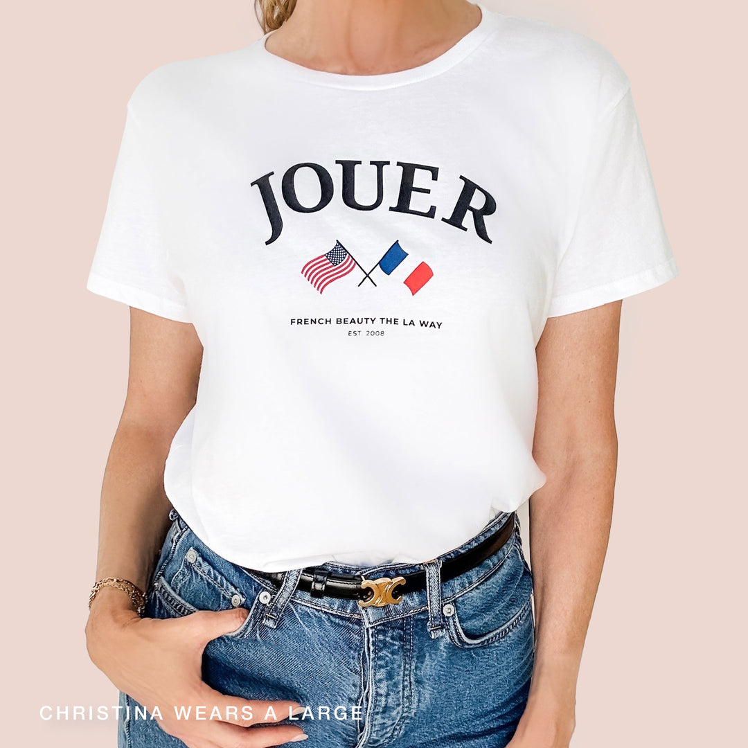 A person is wearing the Paris Meets LA Tee from Jouer Cosmetics, a white 100% cotton t-shirt featuring "JOUER" above an American and a French flag. Below the flags, it says, "FRENCH BEAUTY THE LA WAY EST. 2008." They are also dressed in blue jeans and a black belt. The text at the bottom reads, "CHRISTINA WEARS A LARGE.