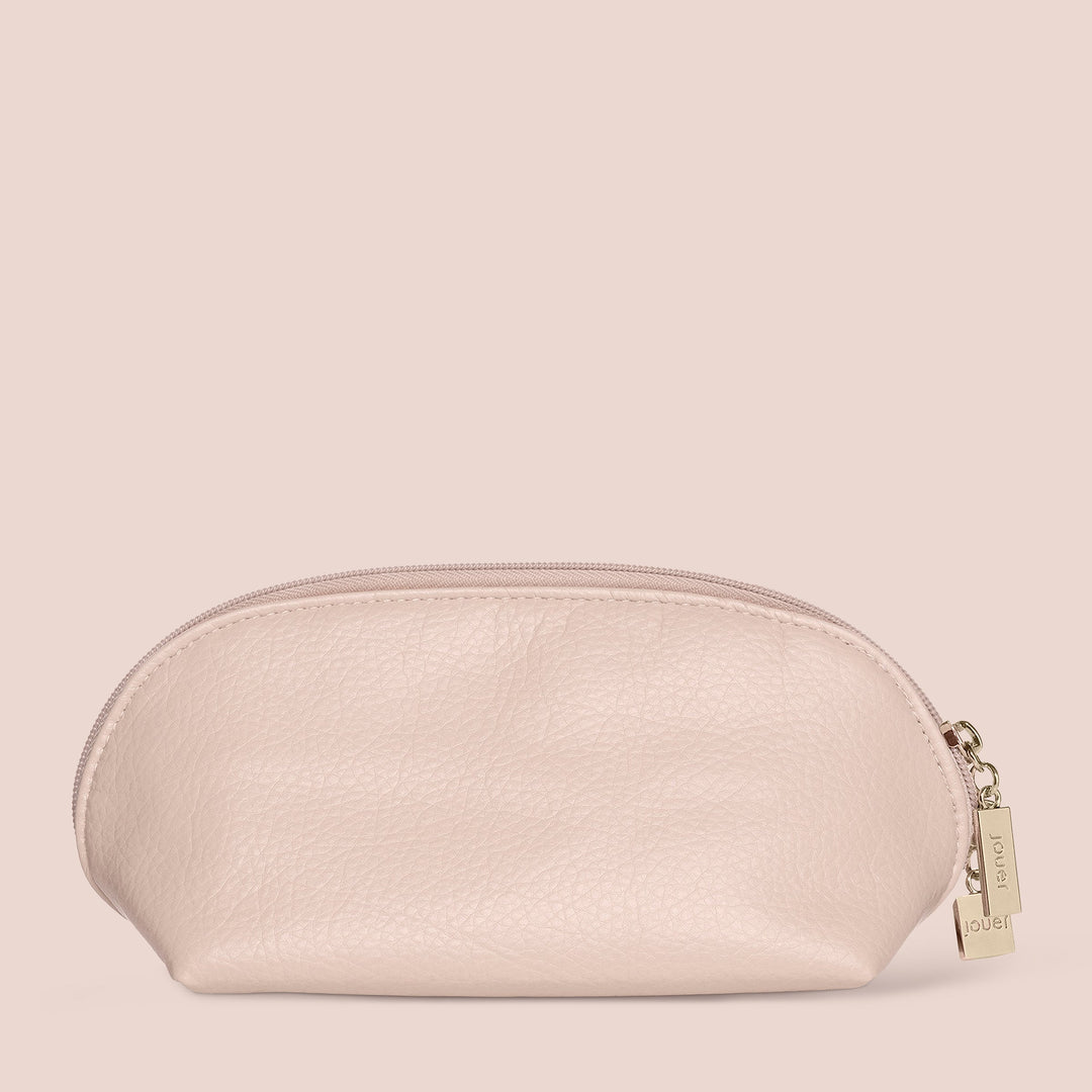 A compact, blush pink leather makeup bag with a zip closure and an elegant gold tag attached to the zipper pull. The dome-shaped makeup bag features a rounded top and flat base, paired against a coordinating pink background. Its travel-friendly design is enhanced by its soft buff leather-like material. This lovely piece is from Jouer Cosmetics' collection.