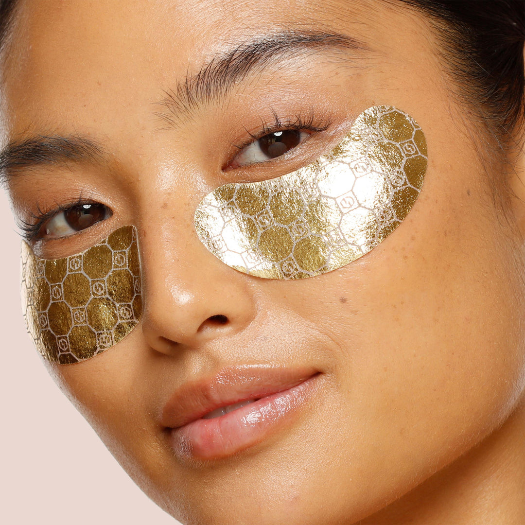 6 pack, A close-up of a person wearing the Jouer Cosmetics *Luminize Eye Patches, which are golden and cruelty-free with a hexagonal pattern. The individual has clear skin, subtle makeup, and is looking directly at the camera with a slight smile. The vegan *Luminize Eye Patches stand out against the plain, light-colored background.