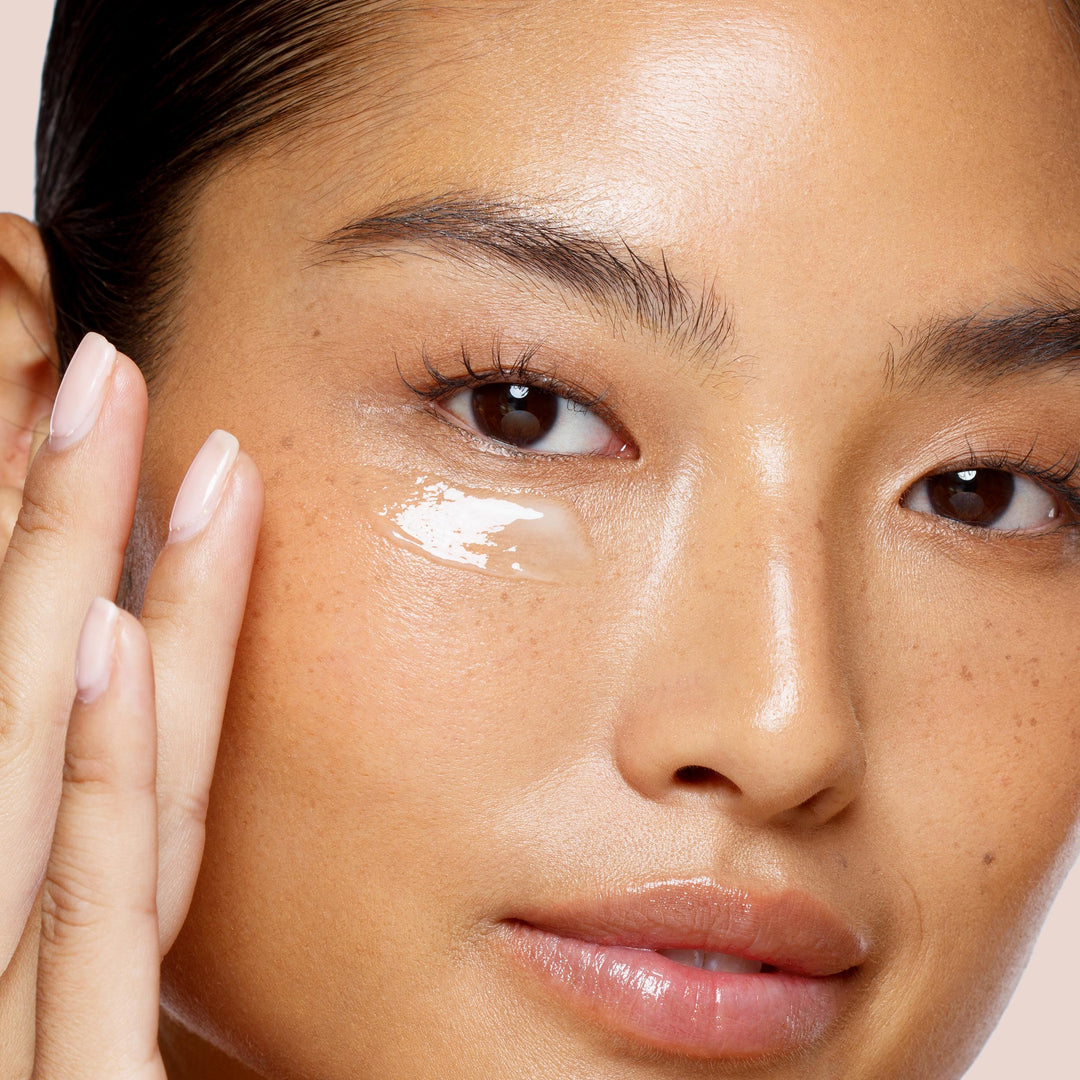 Close-up of a person with smooth, glowing skin applying a small amount of Luminize vegan eye cream from Jouer Cosmetics to the area beneath their right eye using their manicured right index finger. The person's natural, radiant look is enhanced by light makeup.