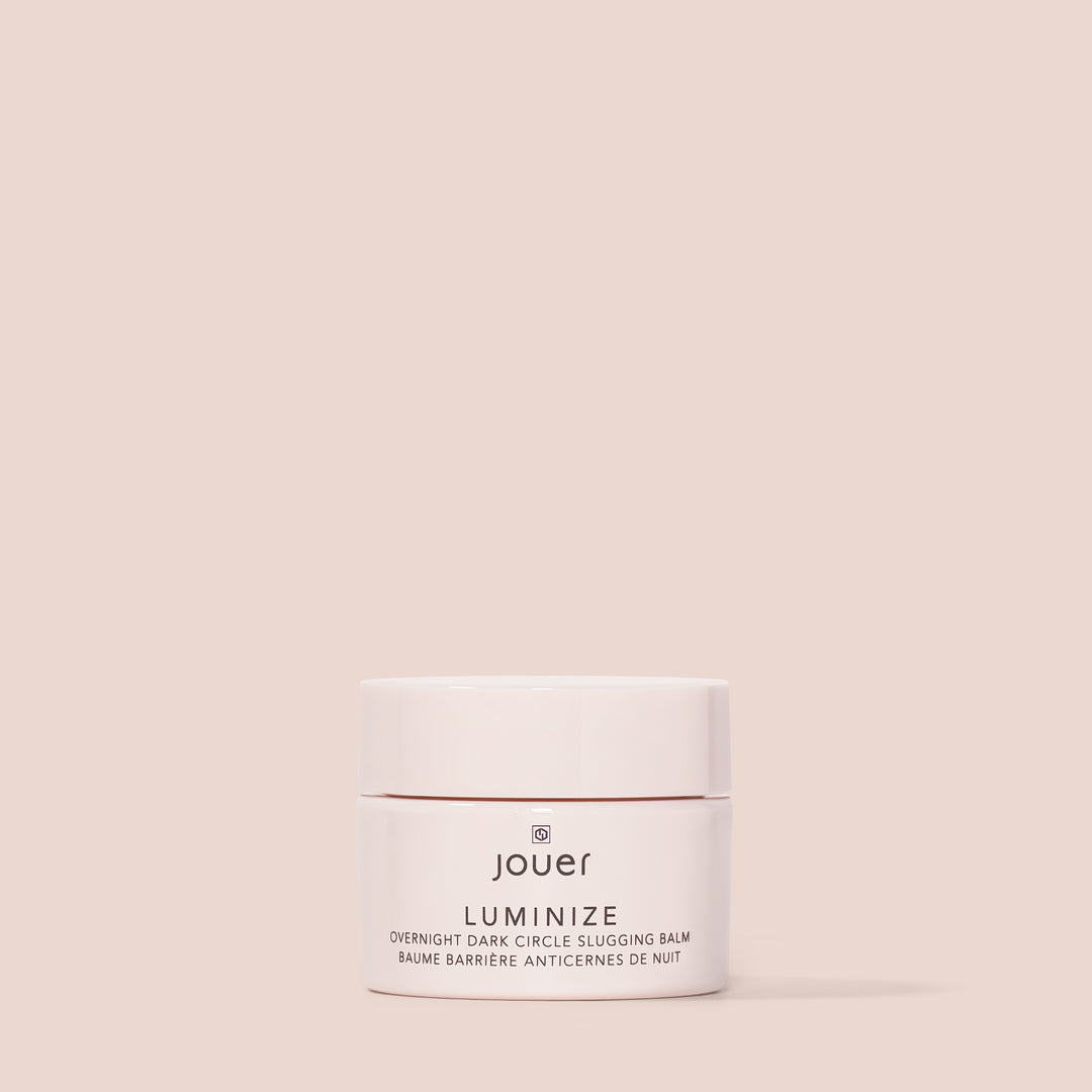 A small, round container of Cruelty-Free Luminize Under Eye Balm by Jouer Cosmetics is centered on a plain beige background. The white and rose gold packaging is minimalist, with the product name "LUMINIZE" prominently displayed on the front.