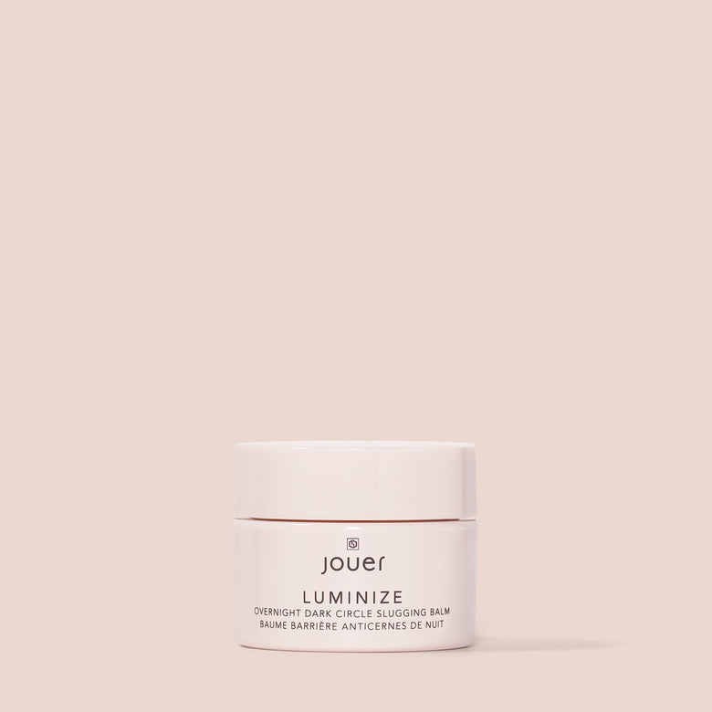 A small, round container of Cruelty-Free Luminize Under Eye Balm by Jouer Cosmetics is centered on a plain beige background. The white and rose gold packaging is minimalist, with the product name 