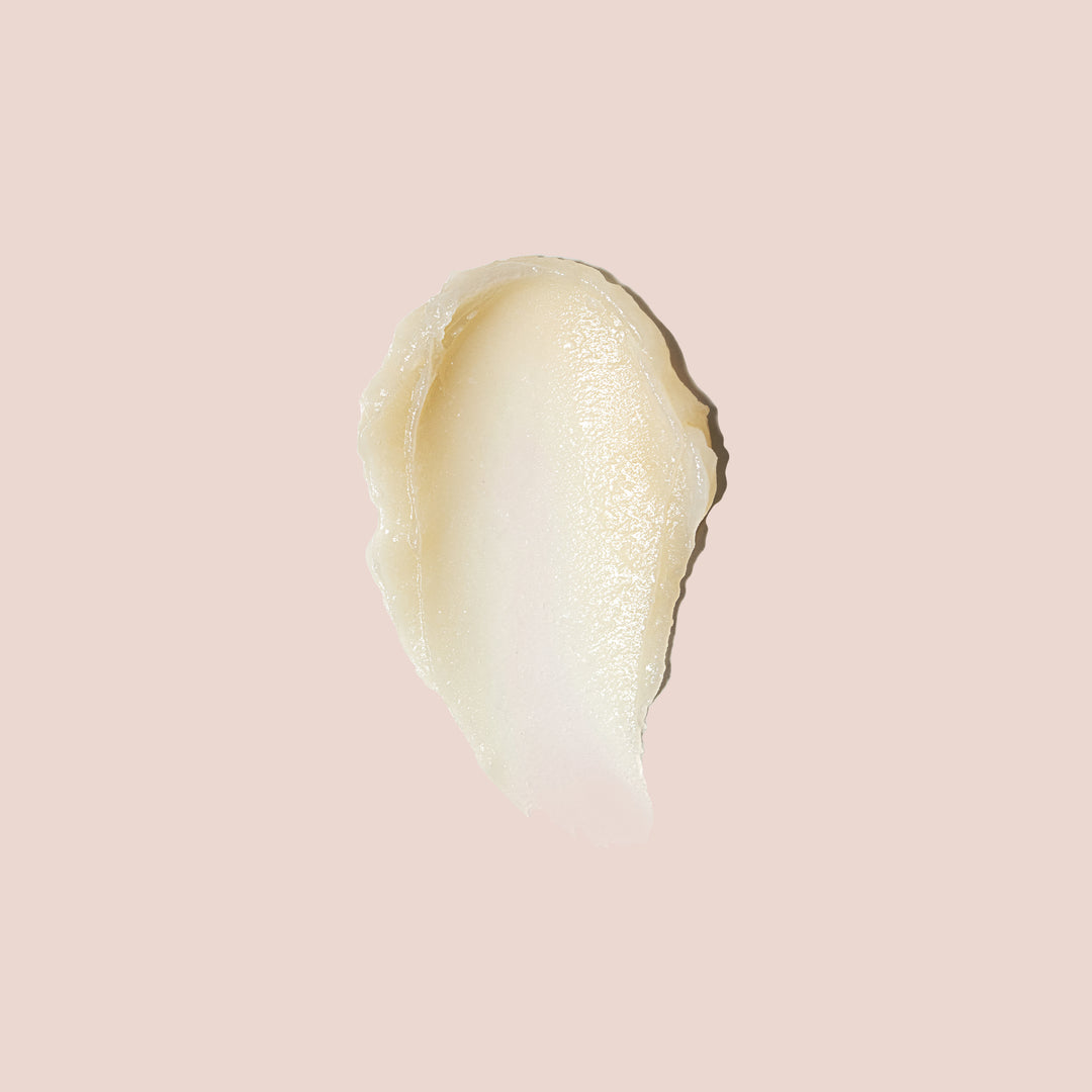 A close-up of a cream-colored, glossy smear of *Luminize Under Eye by Jouer Cosmetics on a solid, light pink background. The texture appears smooth, likely suggesting it is a paraben-free cosmetic product. The smear features irregular edges and a slightly translucent appearance.