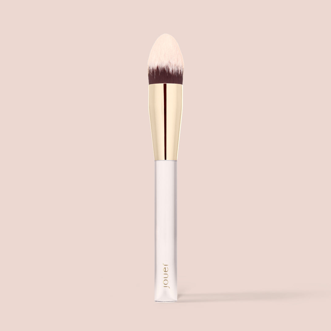 The Essential Concealer Brush by Jouer Cosmetics features a sleek, gold-colored ferrule and a white handle. This vegan and cruelty-free brush boasts soft, tapered bristles that are beige at the tips and darker at the base. The word "jouer" is elegantly printed near the bottom of the handle, all set against a solid light beige background.