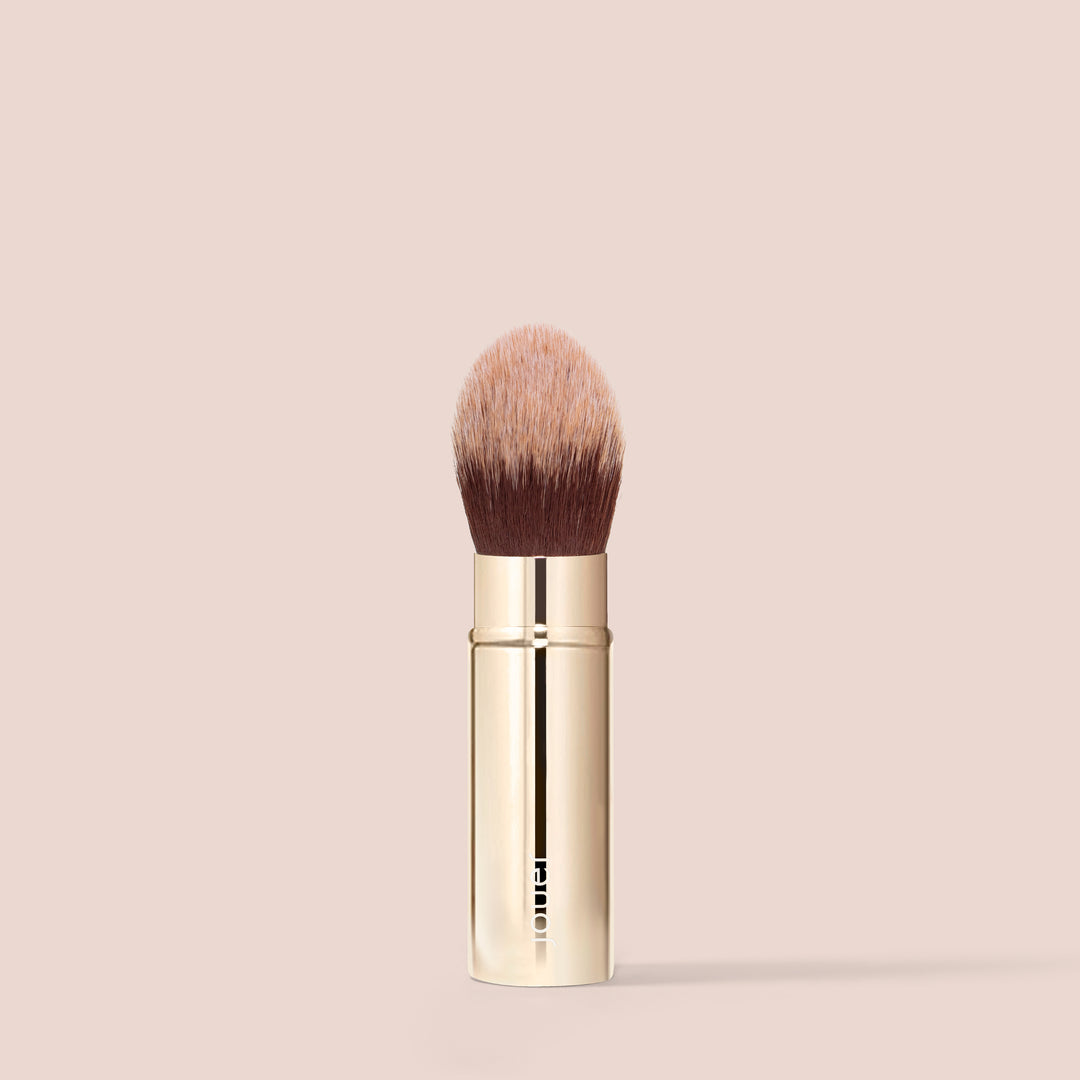 The Essential Complexion Brush by Jouer Cosmetics, featuring soft, round synthetic bristles and a shiny, gold-colored handle, stands upright against a beige background. The vegan, cruelty-free brush appears to be clean and unused.