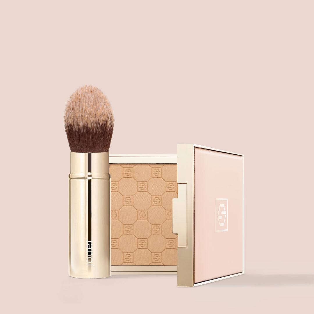 The Essential Complexion Brush from Jouer Cosmetics, featuring a gold handle and soft, synthetic bristles, stands beside an open compact with a hexagon-patterned powder on a beige background. The light pink compact, adorned with gold accents and proudly displaying its logo on the lid, highlights both vegan and cruelty-free products preferred by conscious beauty lovers.