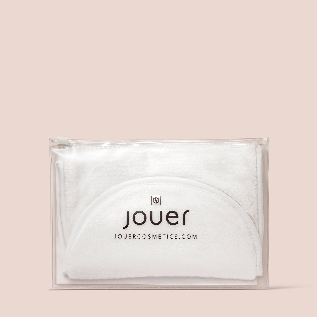 A transparent zippered plastic pouch displaying folded white Microfiber Towels for a gentle wipe to remove makeup. The pouch features the Jouer Cosmetics logo and their website address, jouercomestics.com, printed in black on the front. The background is a neutral beige color.