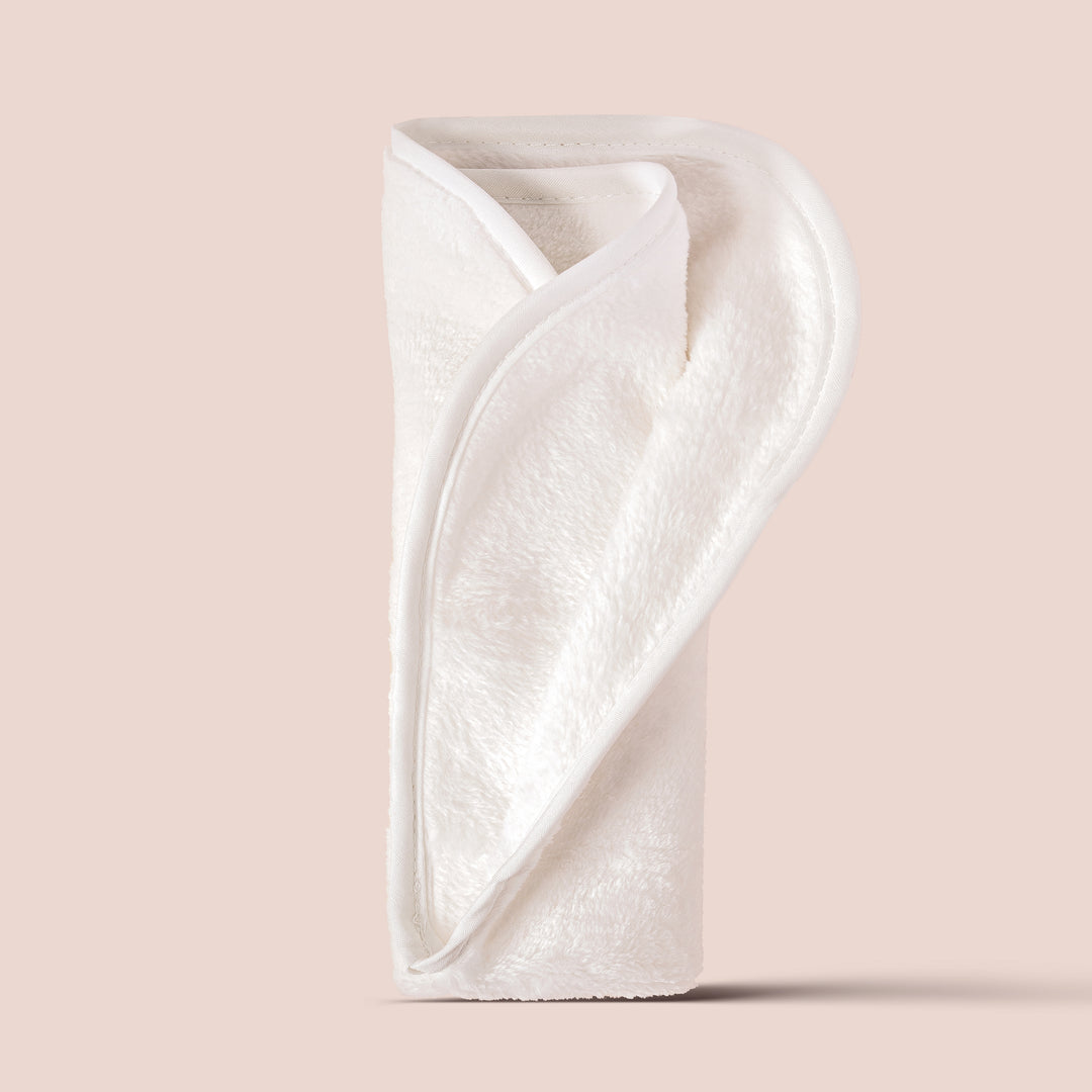 An upright, folded Jouer Cosmetics Microfiber Towel with a neatly arranged overlapping edge, set against a plain beige background. The white towel appears plush and soft, perfect for a gentle wipe to remove makeup. It has a slight sheen on its surface.