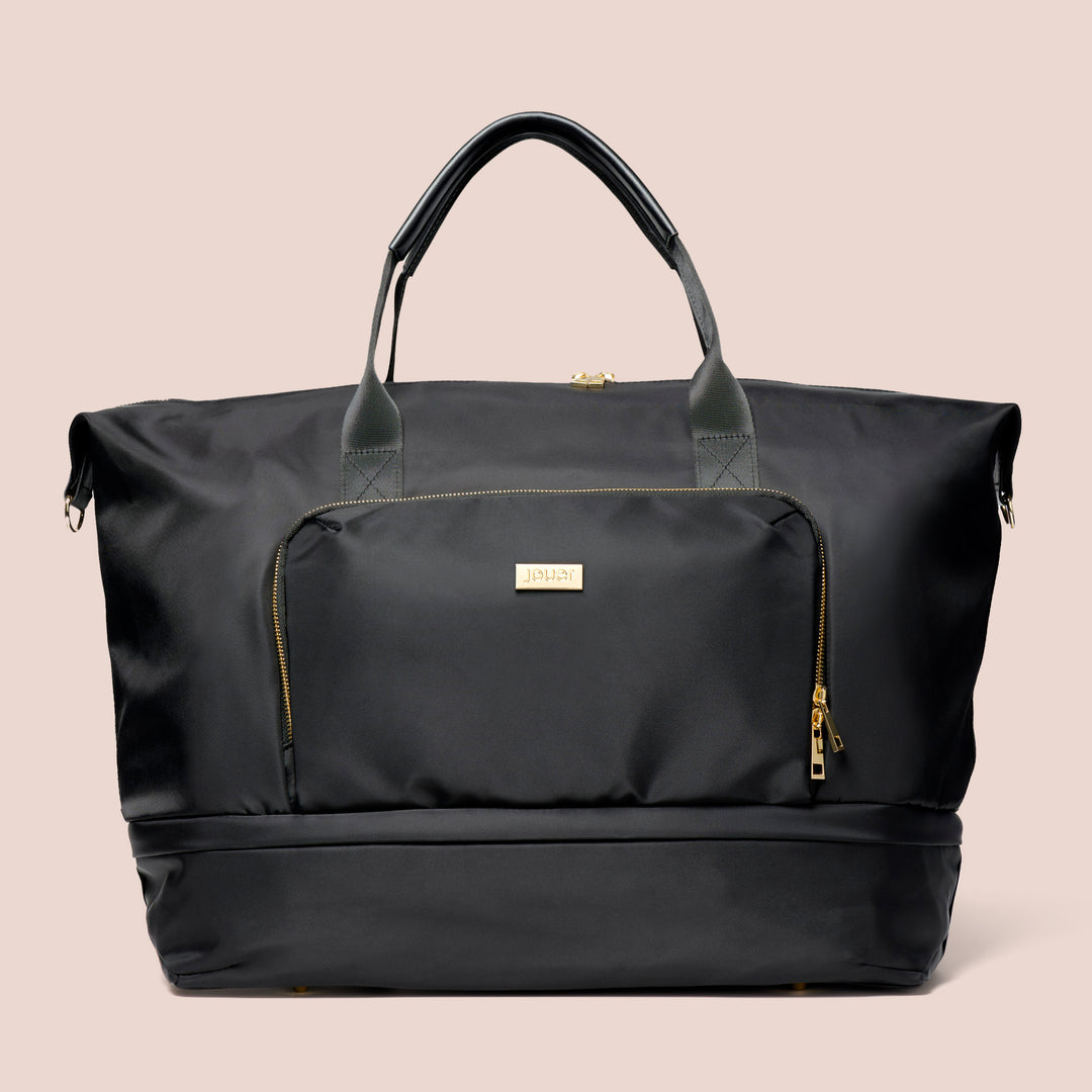 A black Le Weekender Travel Bag by Jouer Cosmetics, showcasing a sleek design, is featured against a plain background. It comes with dual handles, a front zippered pocket, and displays a central logo tag. The bag highlights gold zippers and accents, along with an expanding zippered compartment for additional storage.