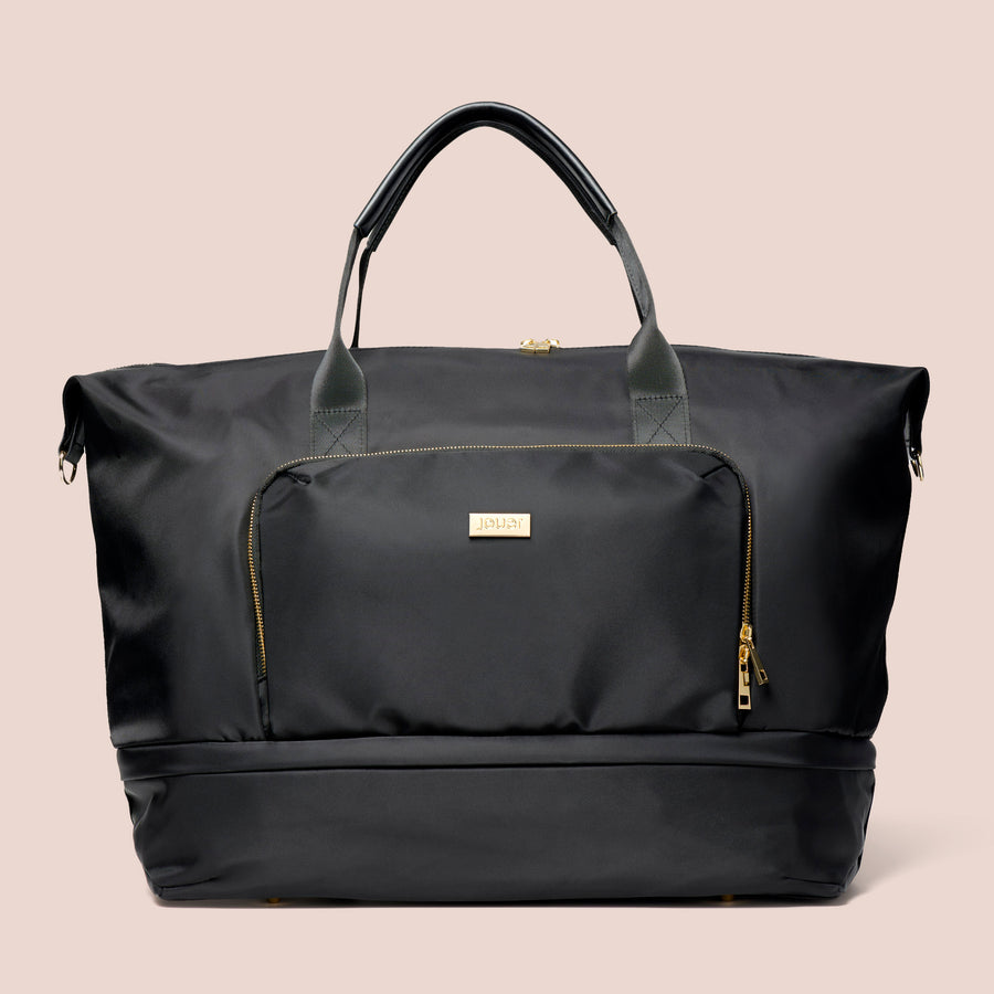 Black Le Weekender travel bag with gold zipper details and front pocket against pink background