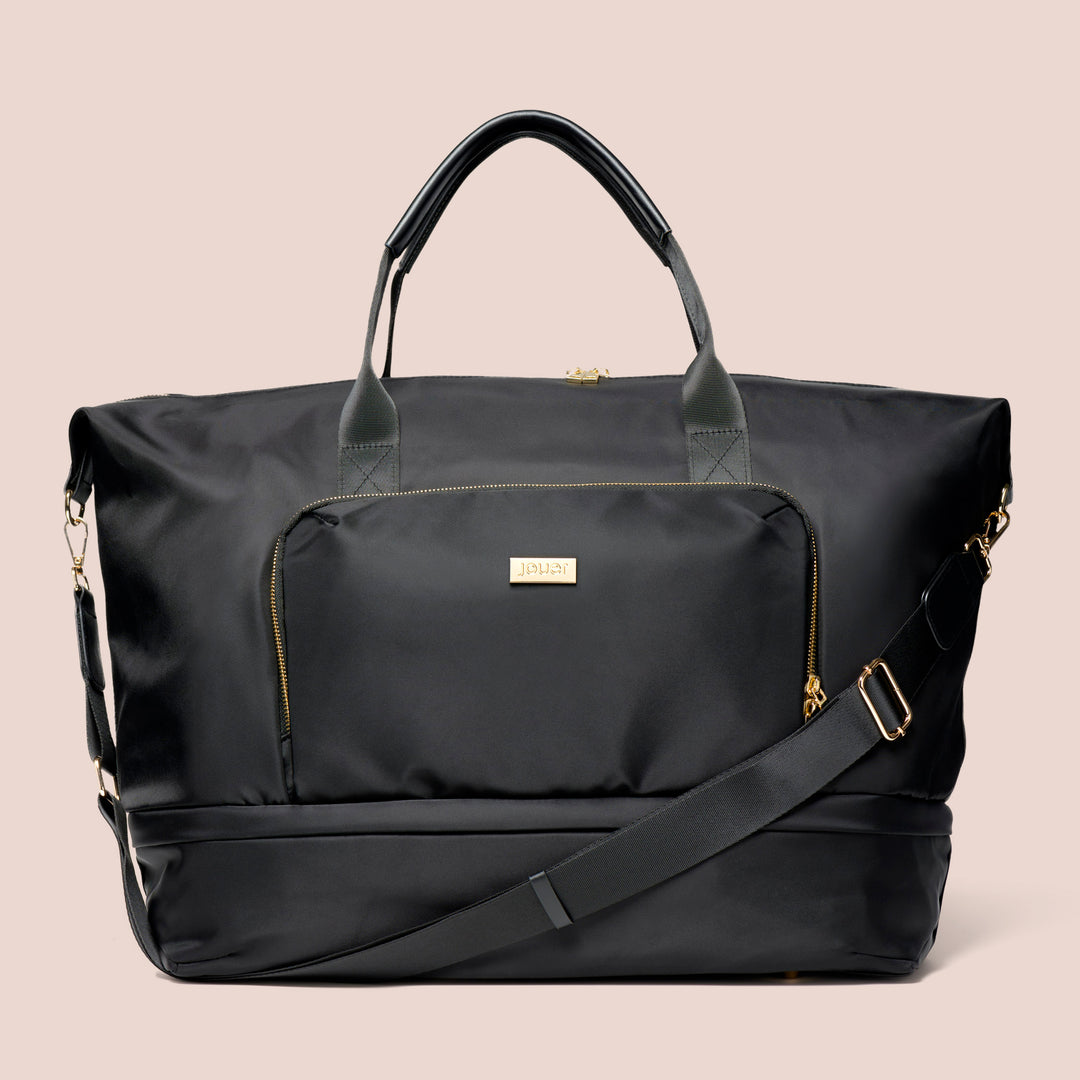 The Le Weekender Travel Bag by Jouer Cosmetics is a sleek black duffel bag with dual leather handles, a removable shoulder strap, and gold-tone hardware. The bag features a zippered exterior pocket adorned with a gold brand label and interior zippered pockets for added organization, all set against a neutral beige background.