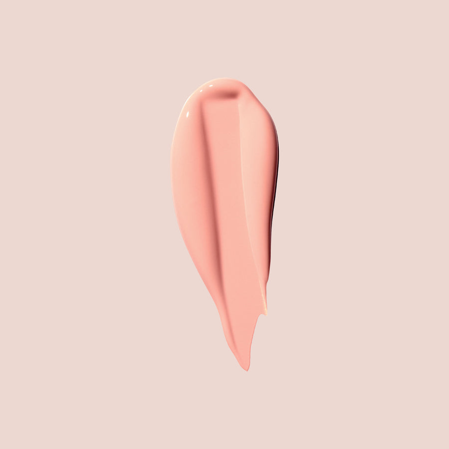 A smooth, glossy swatch of light peachy pink cosmetic under-eye corrector, shaped like a teardrop, centered on a solid pale pink background.