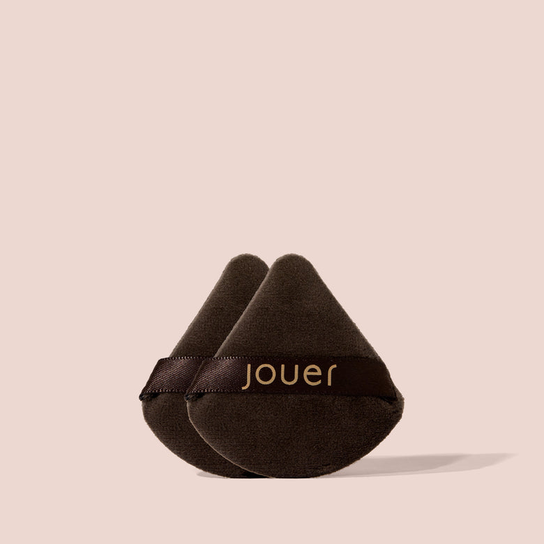Two Jouer Perfecting Puffs in black velvet with gold logo ribbon stacked together on a beige background.