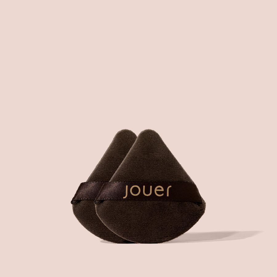Two Jouer Perfecting Puffs in black velvet with gold logo ribbon stacked together on a beige background.