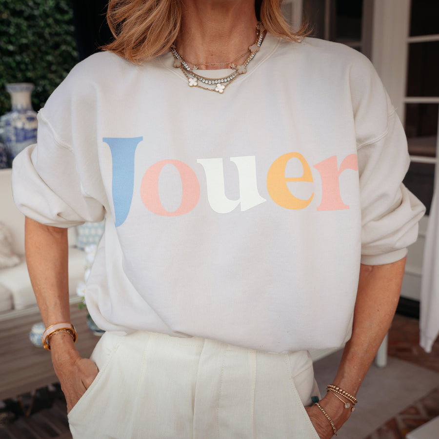 Woman wearing cream Jouer pastel sweatshirt with colorful logo text and layered pearl necklaces
