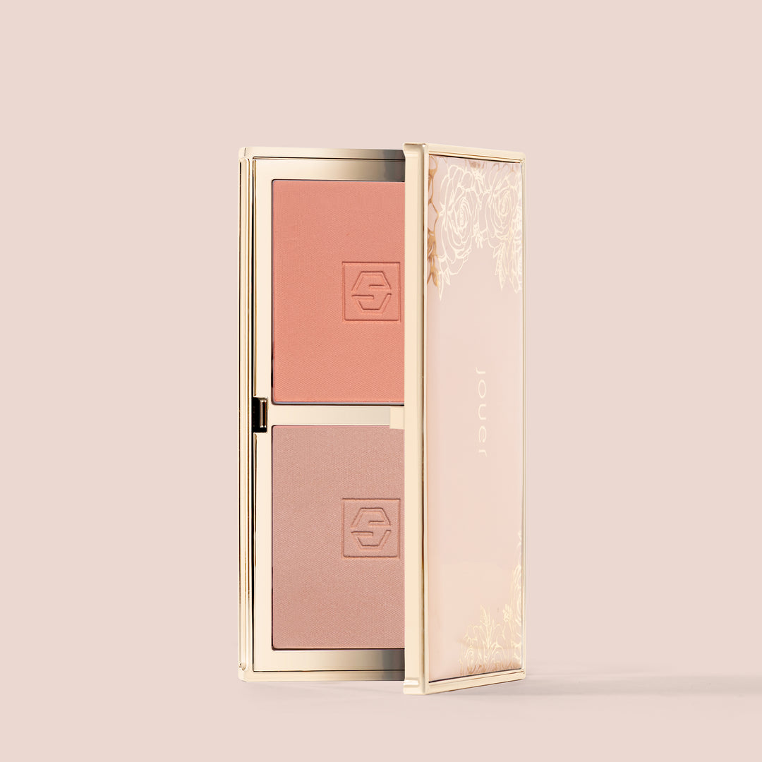 adore, The Blush Bouquet by Jouer Cosmetics is displayed open, showcasing two hues of blush in a side-by-side arrangement. The compact case features a floral design on the lid's exterior and uses a vegan formula. One blush is in a peachy hue, while the other offers a lighter, more nude tone.