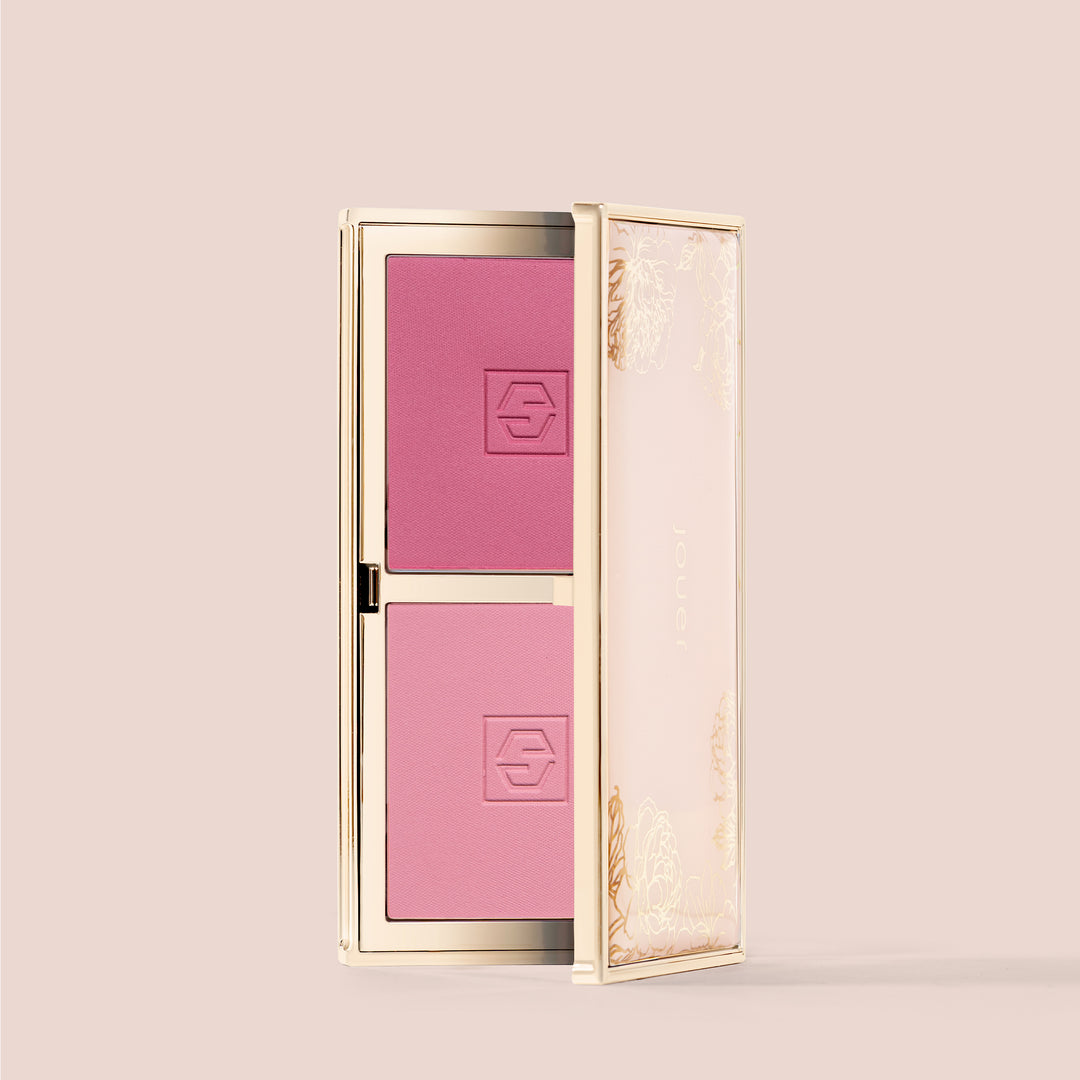 coquette, A sleek golden compact called Blush Bouquet by Jouer Cosmetics contains two shades of pink blush. The compact is slightly open, revealing the blush inside. The packaging features an elegant, floral design. The cruelty-free and vegan formula blush is pressed into square pans with a subtle logo imprint on each.