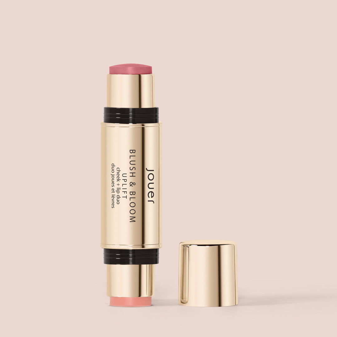 Uplift, The "Blush & Bloom" dual-ended makeup stick by Jouer Cosmetics is a clean, cruelty-free, and vegan product. It features a gold-colored casing with one end displaying a soft pink blush, while the other end is capped and likely contains another makeup item. The cap is detached and placed beside the stick.