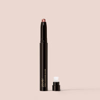 rose gold, A black Crème Eyeshadow from Jouer Cosmetics with a detached cap is standing upright against a beige background. The tip of the paraben-free crayon is a neutral pink shade, and the cap rests nearby, revealing a built-in brush for application.