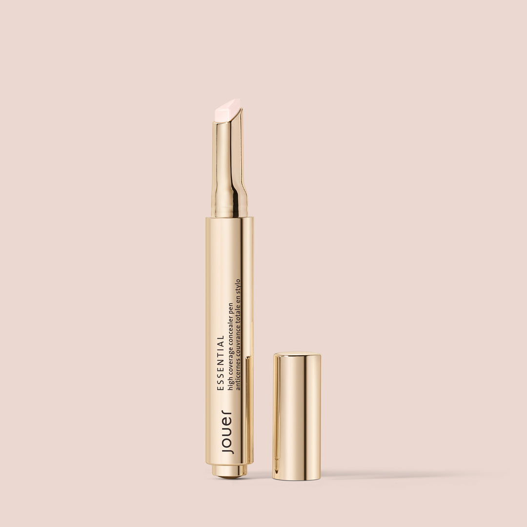 snow, A sleek, gold-colored tube of lip enhancer with the cap removed reveals a light pink, angled lip product. The name "Jouer Cosmetics" and the title "Essential Concealer Pen" are printed on the side of the tube. This vegan beauty essential is set against a solid, soft beige background.