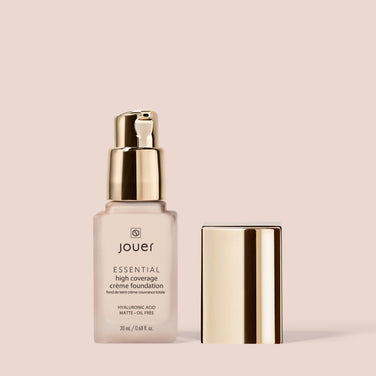  The Essential Crème Foundation by Jouer Cosmetics comes in a light beige bottle with a gold pump dispenser and a matching gold cap placed beside it. The bottle features text stating that the foundation contains hyaluronic acid, offers a matte, oil-free finish, and is cruelty-free, making it an ethical choice in beauty.