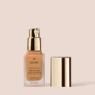 A beige bottle of Jouer Cosmetics Essential Crème Foundation with a gold pump dispenser is shown on a neutral background. The product label highlights features such as hyaluronic acid, matte finish, and an oil-free formula. This cruelty-free product also has a gold cap placed beside it.
