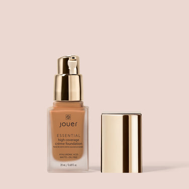  bottle of Jouer Cosmetics Essential Crème Foundation with a gold pump dispenser and a matching gold cap placed next to it. The bottle displays product details, including "Hyaluronic Acid," "Matte+Oil Free," and "Gluten Free" on a beige background.