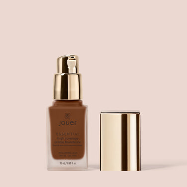  A sleek bottle of Jouer Cosmetics Essential Crème Foundation with a gold pump dispenser and cap is shown against a light beige background. The cruelty-free foundation bottle, labeled with product details, contains 20 ml (0.68 fl oz) of liquid foundation in a rich brown shade.