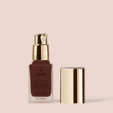  A medium brown bottle of Jouer Cosmetics Essential Crème Foundation, boasting a Paraben Free and Cruelty Free formula, comes with a gold pump dispenser and lid. The bottle contains 20 ml of the product, with the lid positioned separately beside it against a light beige background.